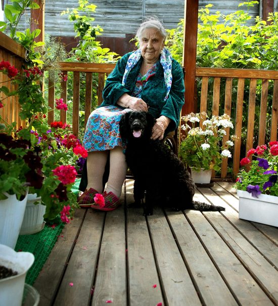 Senior woman sitting with her dog on a colorful garden porch, surrounded by flowers.